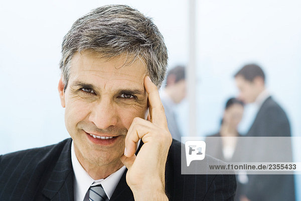 Businessman holding head  smiling at camera  colleagues in background