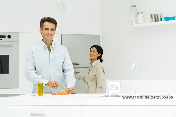 Mann und Frau in der Küche  Mann hackt Tomaten  während Frau in Richtung Kühlschrank geht  beide lächeln in die Kamera.