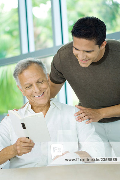 Mature man reading book  adult son looking over his shoulder  both smiling