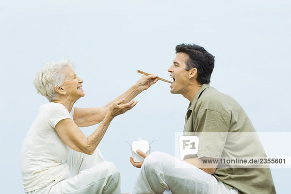Seniorin füttert erwachsenen Sohn mit Stäbchen  lachend  Seitenansicht