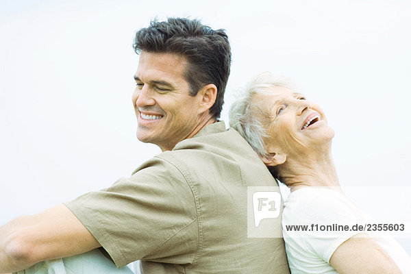 Seniorin und erwachsener Sohn sitzen Rücken an Rücken  lächelnd  Seitenansicht