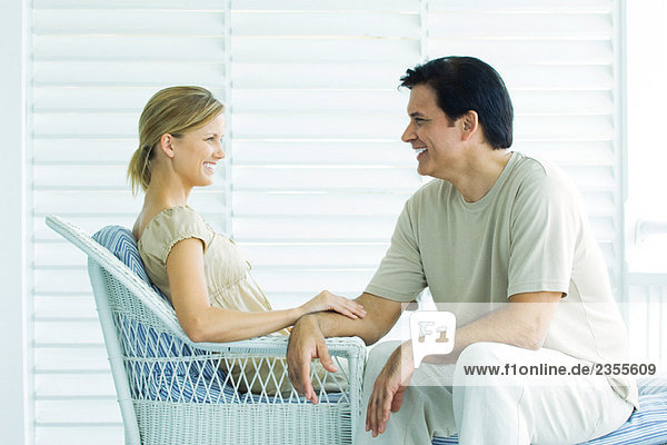 Paar sitzt von Angesicht zu Angesicht auf der Veranda  lächelnd  Frau berührt den Arm des Mannes.