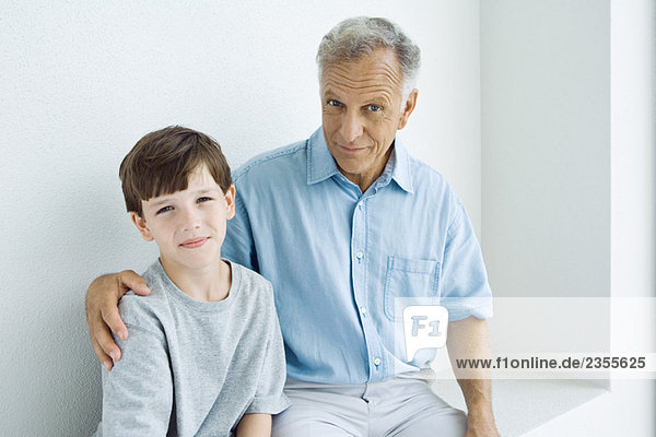Grandfather sitting with arm around grandson's shoulder  both smiling at camera  portrait