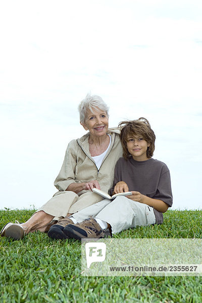 Großmutter und Enkel sitzen im Gras mit Buch  schauen weg und lächeln.