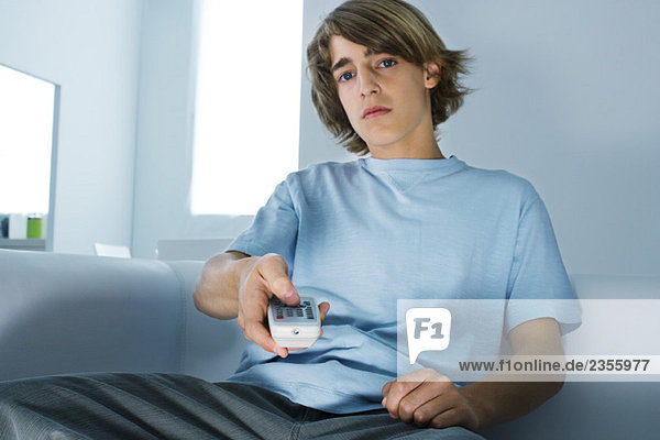 Teenager-Junge sitzt auf der Couch  hält die Fernbedienung und schaut in die Kamera.