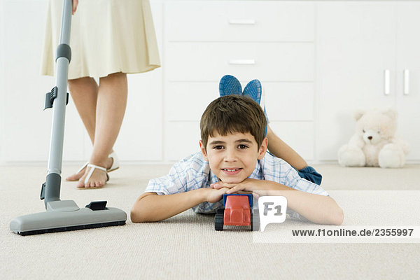 Kleiner Junge mit Spielzeug auf dem Boden liegend  lächelnd in die Kamera  Mutter staubsaugend um ihn herum.