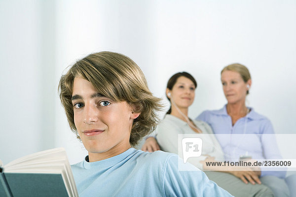 Teenage boy holding book  smiling at camera  women listening to MP3 player in background