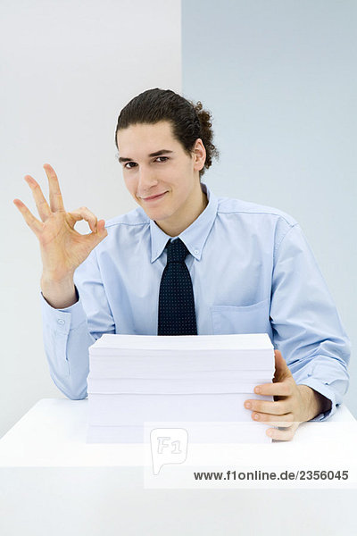 Young professional sitting behind stack of papers  making OK hand gesture  portrait