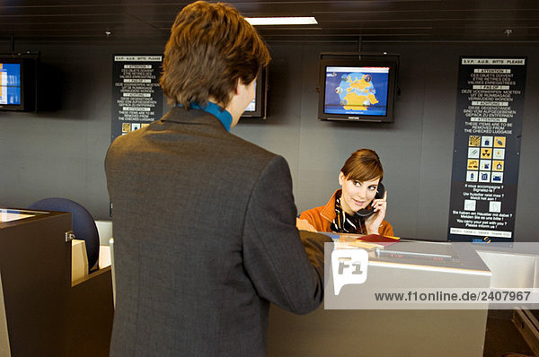 Rear view of a businessman standing at an airport check-in counter and the check-in attendant talking on the telephone