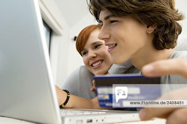 Close-up of a teenage boy holding a credit card and using a laptop with a young woman sitting beside him