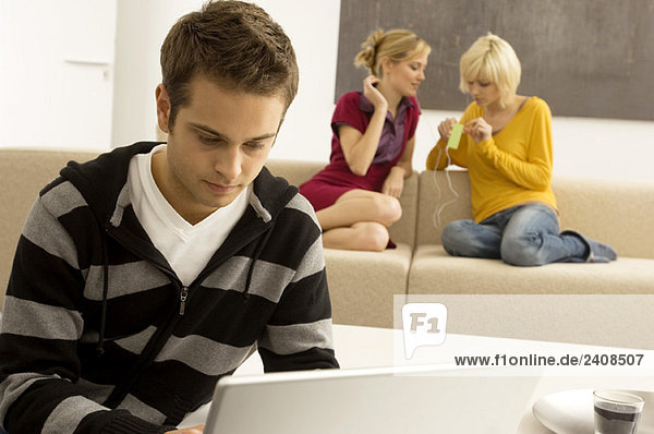 Junger Mann mit einem Laptop und zwei jungen Frauen  die im Hintergrund einen MP3-Player hören.