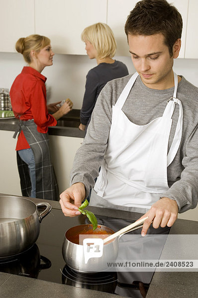Junger Mann bei der Zubereitung des Essens mit zwei jungen Frauen im Hintergrund.