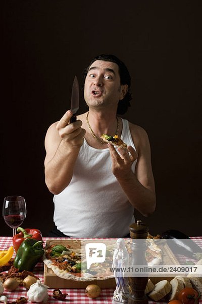 Stereotypischer italienischer Mann  der Pizza isst und ein Messer in der Hand hält.