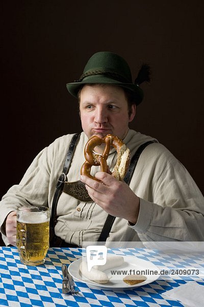Stereotypischer deutscher Mann in bayerischer Tracht  der eine Brezel isst und ein Bier trinkt.