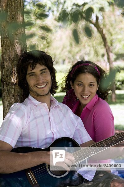 Ein junges Paar sitzt unter einem Baum und spielt Gitarre.