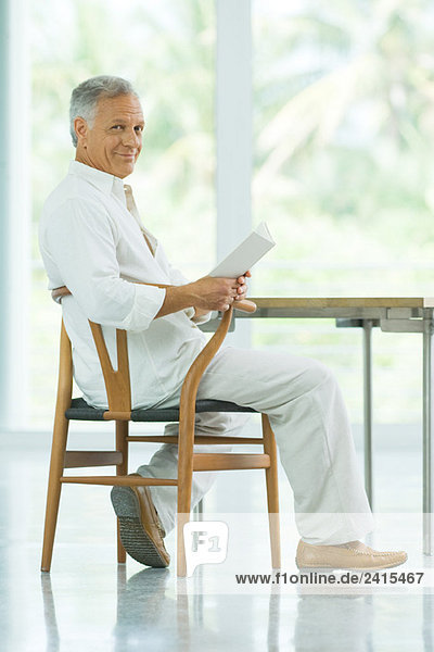 Mature man sitting at table  holding book  smiling at camera