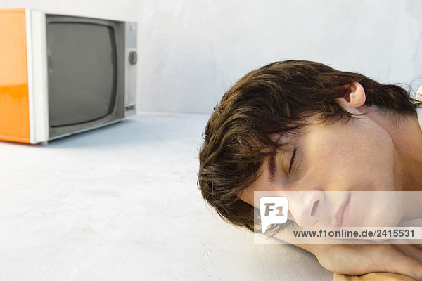 Junger Mann ruht Kopf auf Armen  Augen geschlossen  Fernseher im Hintergrund