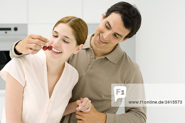 Young couple in kitchen  man holding cherries in front of woman's mouth