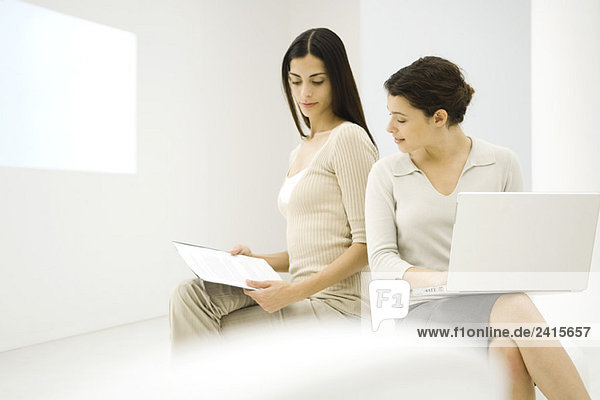 Two female professionals sitting together  discussing document  one holding laptop computer