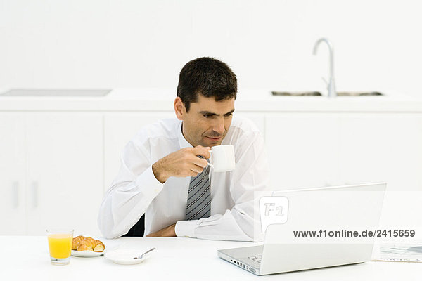 Businessman sitting at kitchen table  looking at laptop computer  drinking coffee