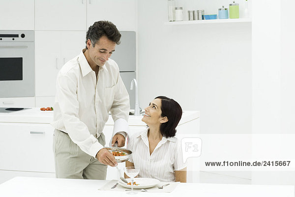 Paar am Küchentisch  Mann serviert Frauenessen  beide lächelnd