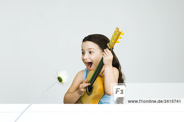 Kleines Mädchen spielt Gitarre und macht ein Gesicht  während sich die Blume zu ihr neigt.