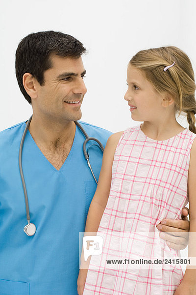 Arzt mit Arm um die Taille des kleinen Mädchens  beide lächeln einander an.