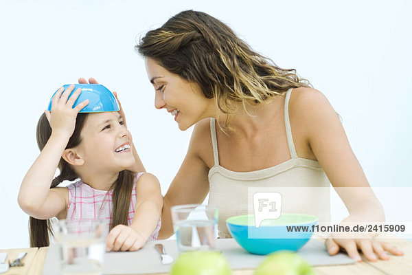 Mutter und Tochter sitzen am Tisch  Mädchen stellt Schale auf den Kopf  beide lächelnd