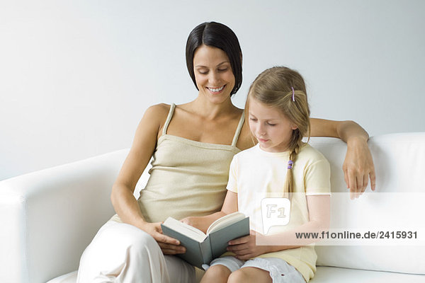 Mutter und Tochter sitzen auf dem Sofa und lesen gemeinsam ein Buch.