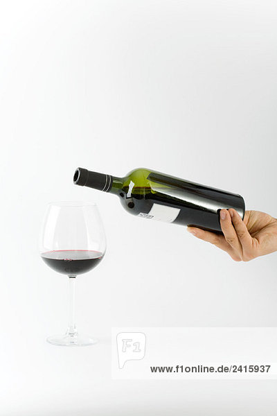 Handgehaltene Weinflasche über Glas mit einer kleinen Menge Rotwein darin