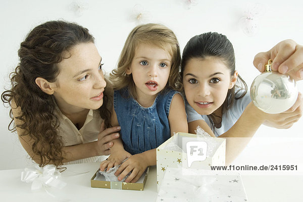 Girl beside mother and sister  holding up Christmas ornament  smiling at camera