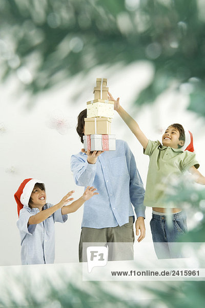 Mann hält Stapel von Weihnachtsgeschenken  zwei Jungen erreichen  gesehen durch Weihnachtsbaumzweige