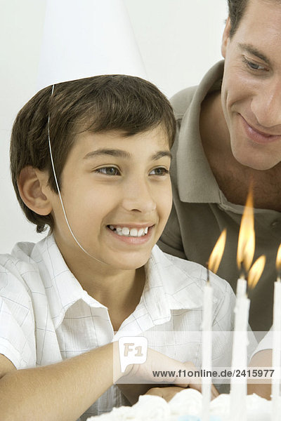 Boy next to birthday cake with lit candles  wearing party hat  smiling