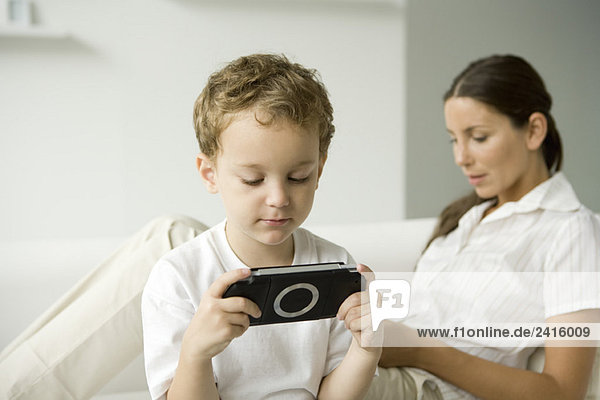 Kleiner Junge spielt Handheld-Videospiel  seine Mutter sitzt auf dem Sofa hinter ihm.