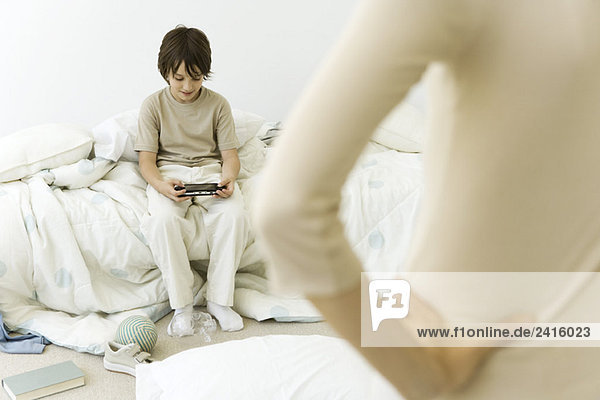 Junge sitzt im unordentlichen Raum  spielt Handheld-Videospiel  Mutter im Vordergrund mit der Hand auf der Hüfte  abgeschnittene Ansicht
