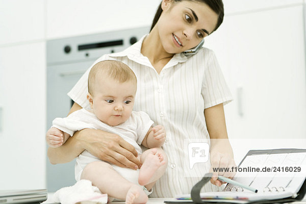 Woman holding baby  using cell phone and looking at agenda