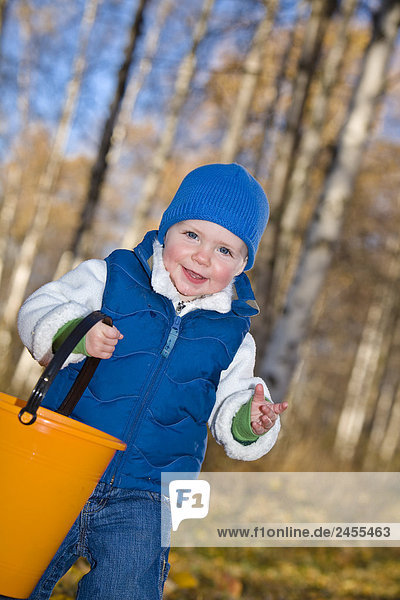 Young girl toddler playing in the Fall leaves carring a orange bucket in a forested area of Anchorage in Southcentral Alaska