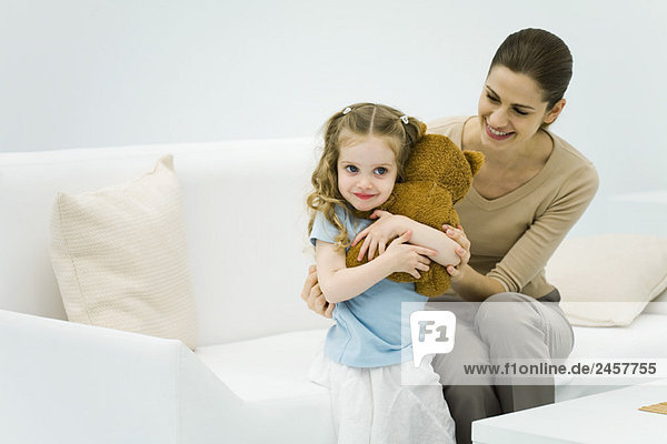 Kleines Mädchen mit Plüschbär  Frau auf der Couch sitzend  kleines Mädchen sanft umarmend