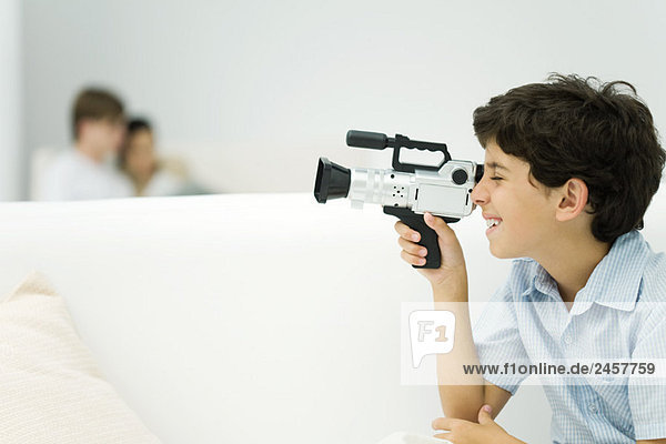 Junge schaut durch Videokamera  Paar im Hintergrund  Seitenansicht