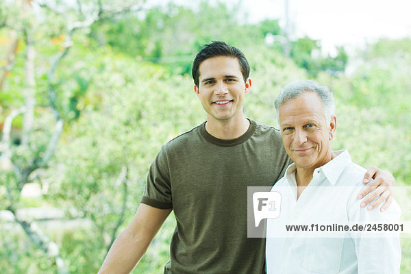 Young man with arm around his father's shoulder  both smiling at camera  portrait