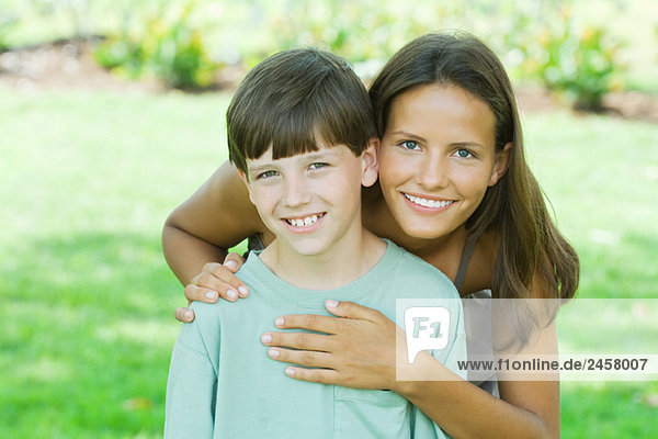 Teen girl standing behind younger brother  hands on shoulder and heart  both smiling at camera