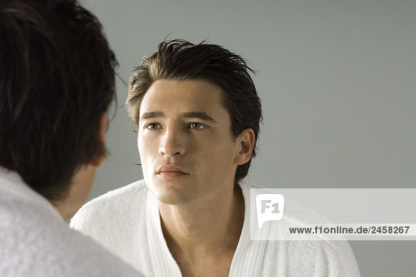 Ein Mann  der sein Gesicht im Spiegel betrachtet und einen Bademantel trägt.