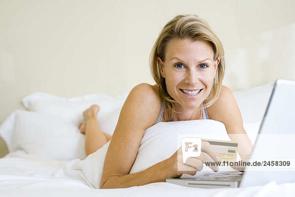 Frau im Bett liegend  mit Laptop  Kreditkarte haltend  Kamera lächelnd