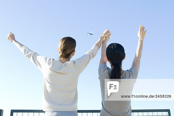 Zwei Frauen  die das Flugzeug am Himmel betrachten  Hände haltend  Arme erhoben  Rückansicht