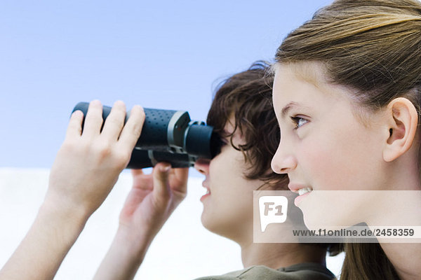Teenage boy looking away through binoculars  sister nearby  profile