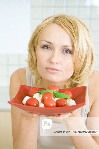 Frau hält einen Teller mit Caprese-Salat hoch und schaut in die Kamera.