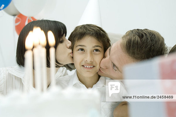 Familie hinter Geburtstagskuchen mit angezündeten Kerzen  Eltern küssen Jungen auf Wangen