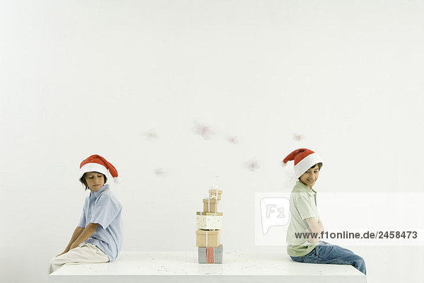 Zwei Brüder sitzen Rücken an Rücken  Weihnachtsgeschenke zwischen ihnen gestapelt  beide mit Weihnachtsmützen.