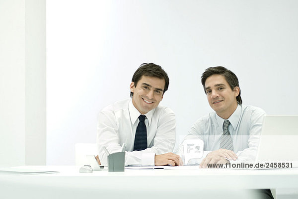 Männliche Geschäftspartner sitzen Seite an Seite am Schreibtisch und lächeln in die Kamera.