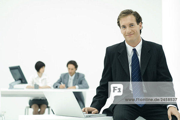 Businessman sitting with laptop  smiling at camera  colleagues in background